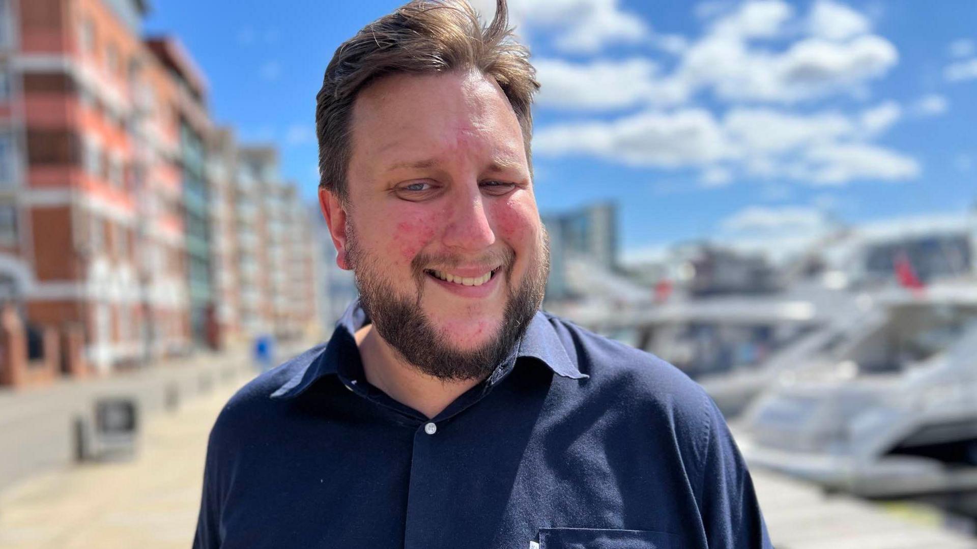 Lee Walker who has dark brown hair and dark brown beard smiling at camera, standing outside against a blurred background of white cruiser boats on the right and office/flat buildings on the left 