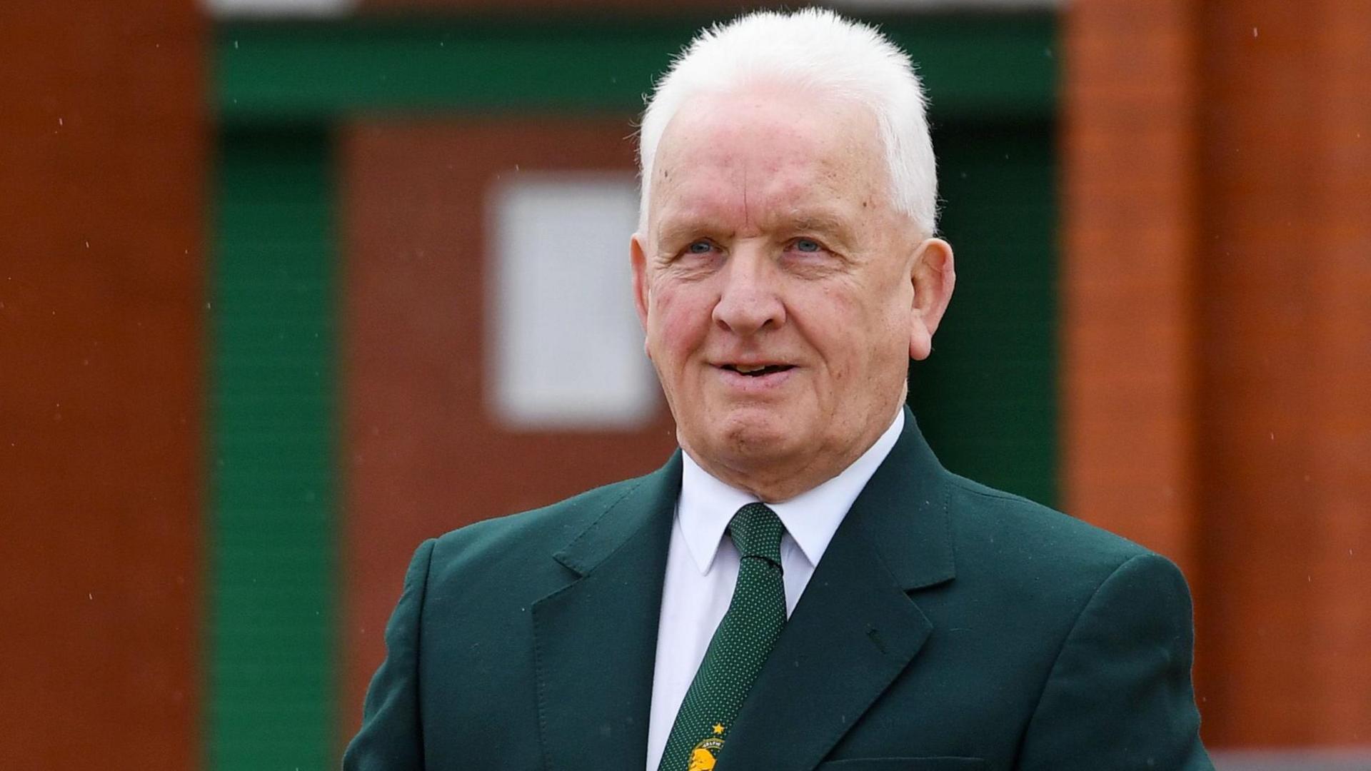 A man with white hair and a green suit jacket and tie gives a small smile to the camera
