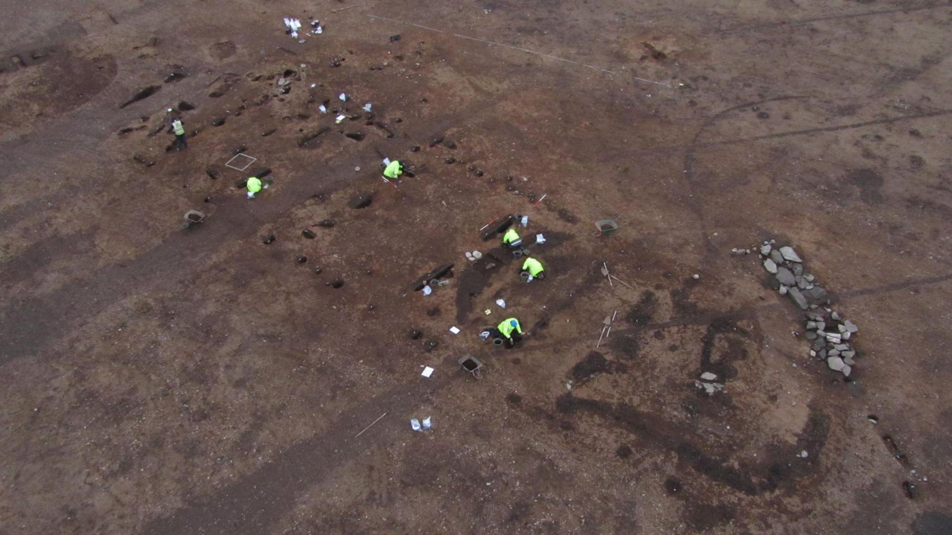 An aerial image of the dig site. The ground is bare, brown earth and archaeologists wearing fluorescent jackets work at uncovering finds. The long "footprint" of the hall is evident.