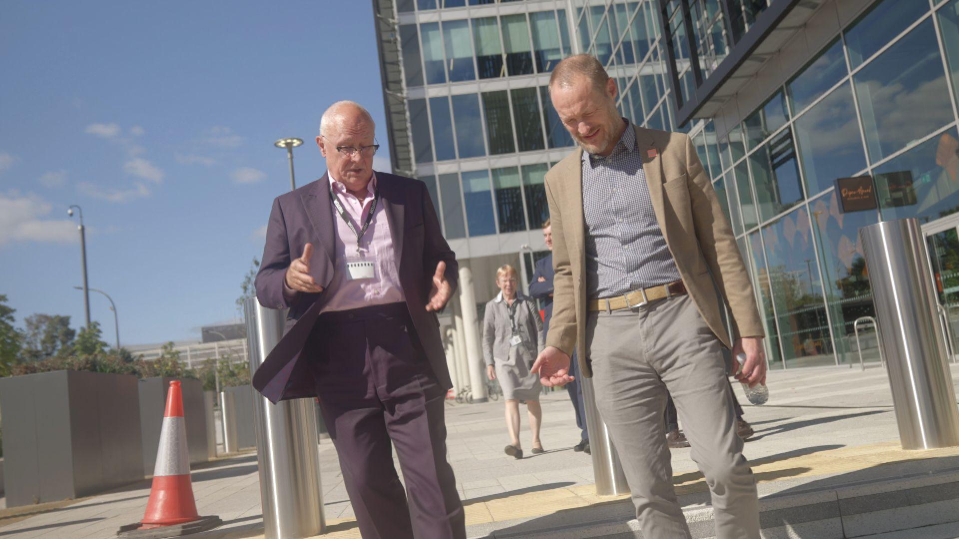 A candid shot of Sir Michael Lyons, wearing a purple suit and pink shirt, in conversation with another man during a tour of Milton Keynes. The other man is wearing a light brown jacket, blue checked shirt and light grey trousers. They both appear to be walking towards the camera. In the background is a glass-panelled office building. The weather appears fine and a patch of blue sky can be seen.