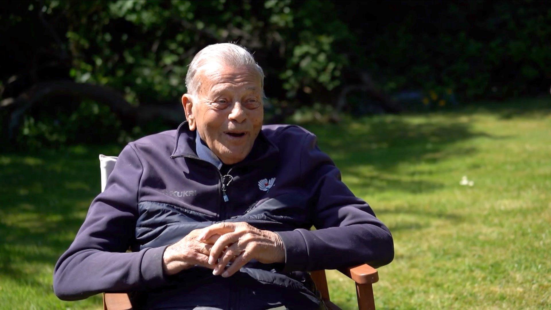 Dickie Bird sitting down in a wooden chair in his garden, smiling in the sun.