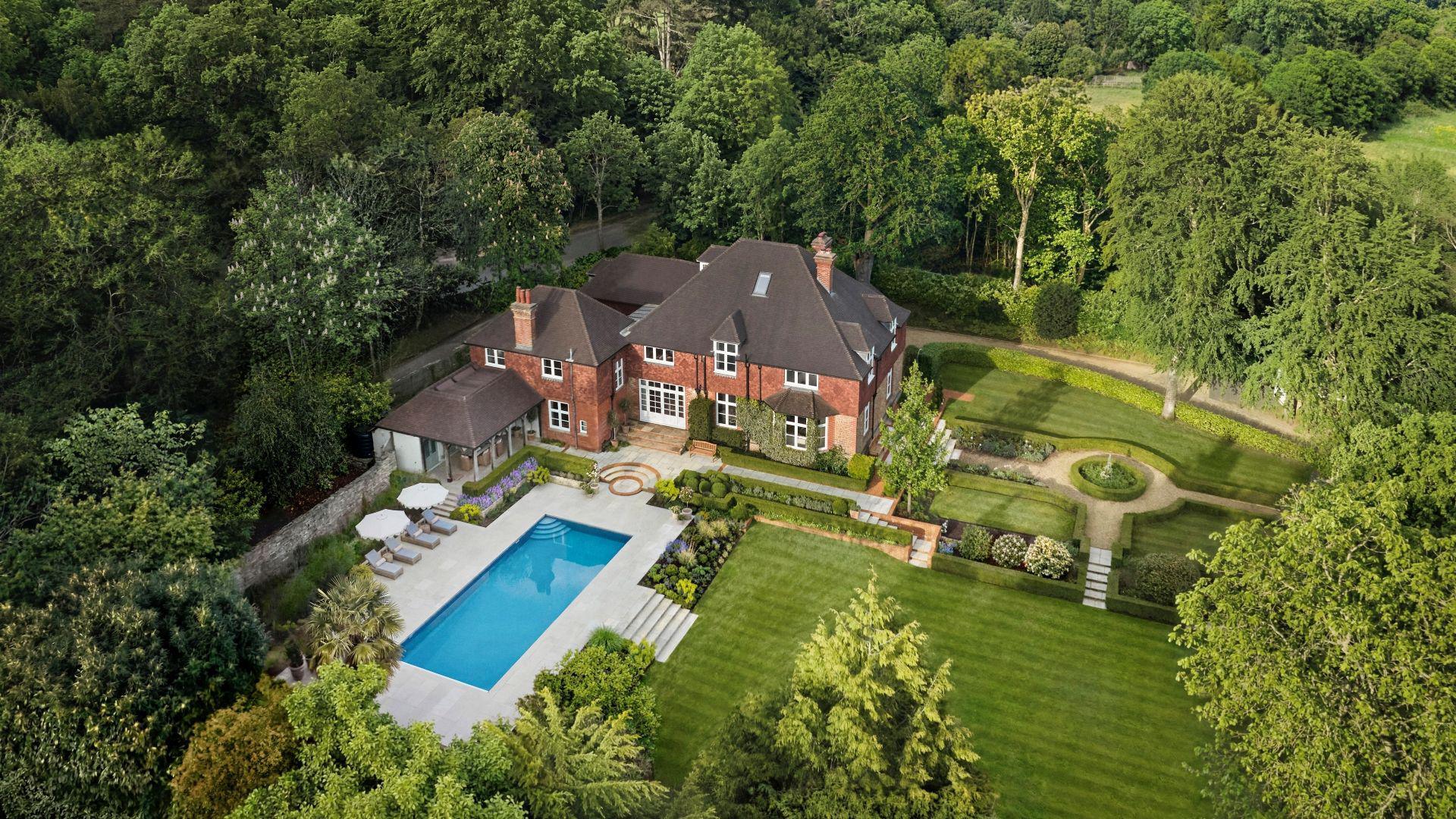 An aerial shot of a large house with a swimming pool set within a manicured garden and surrounded by trees