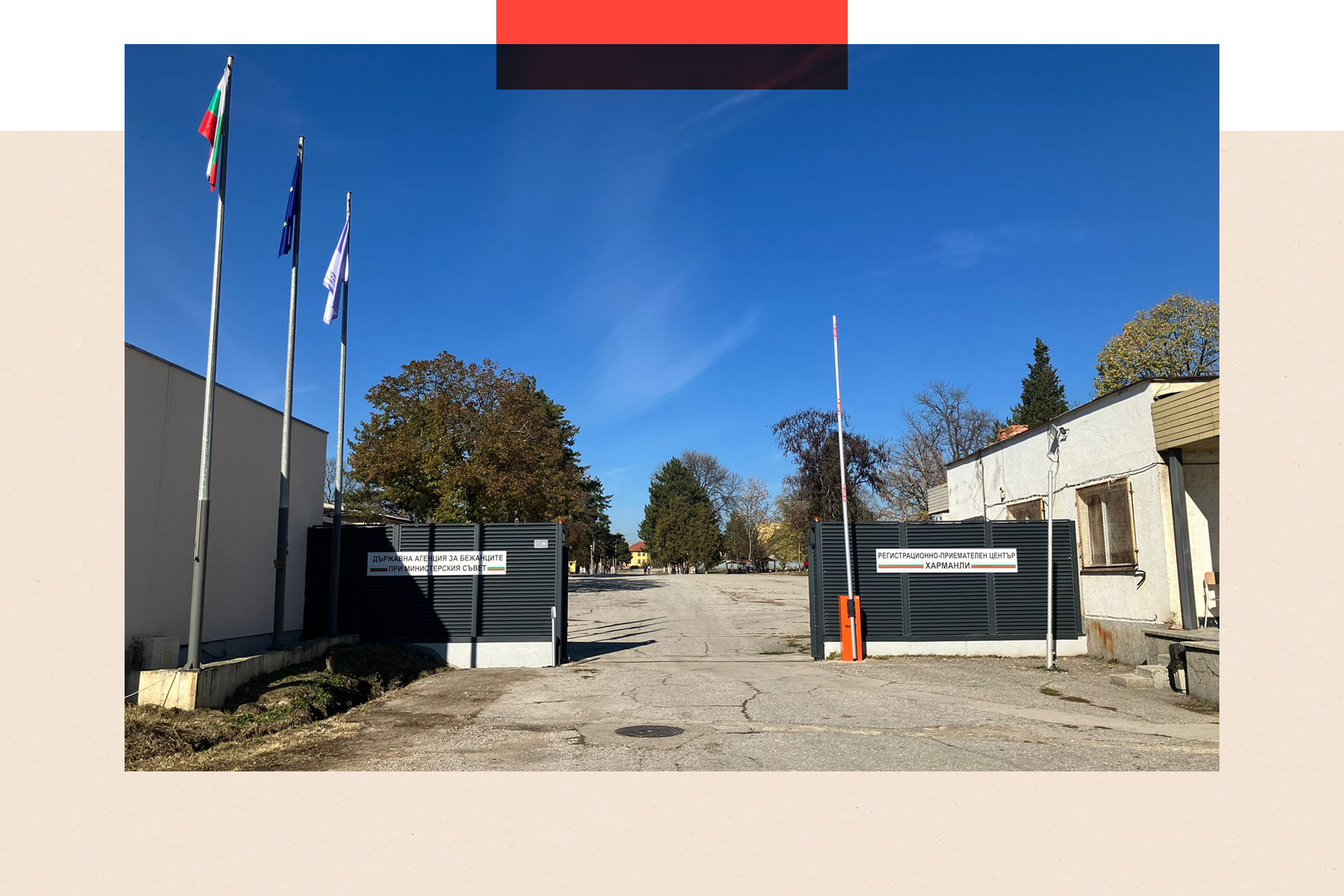 Photo of the exterior of the Harmanli refugee camp. The main gate is open with the car barrier raised and a concrete expanse behind. There are trees and the sky is blue.