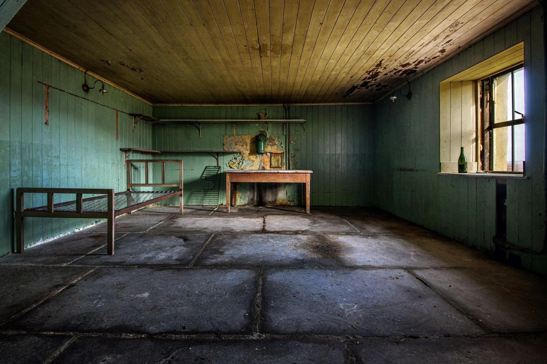 The room has a flagstone floor. There is a bed frame and wooden table. Light comes in from a window.