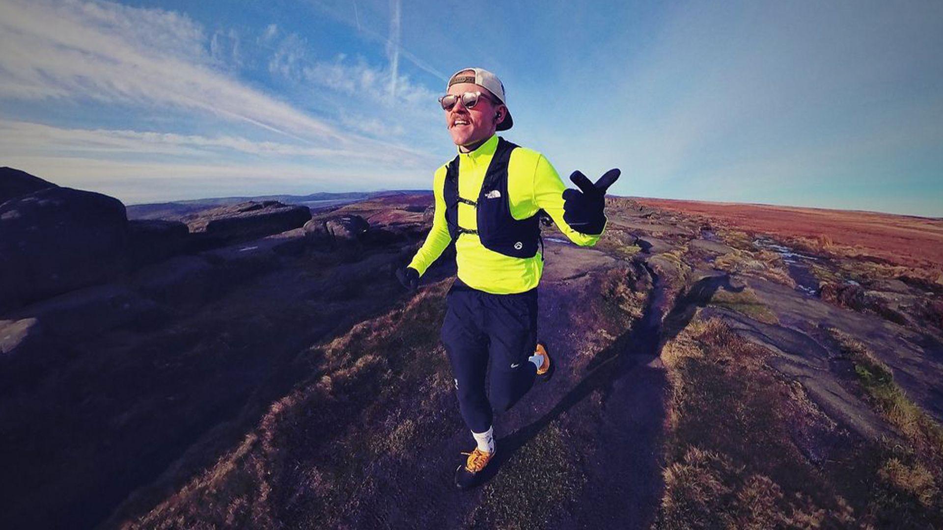 Andy Hobson dressed in high-vis running gear on rolling hills. He is pointing to the camera and wearing sunglasses.