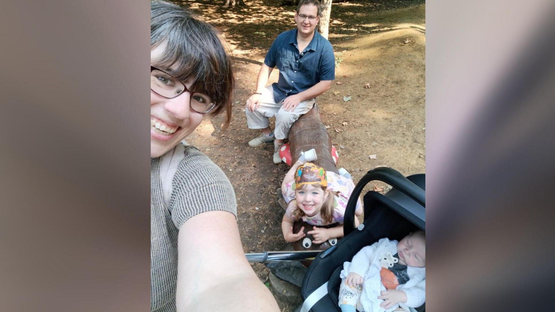 Selfie of Adele holding the phone - Oliver in a buggy, her daughter and husband sitting on a log