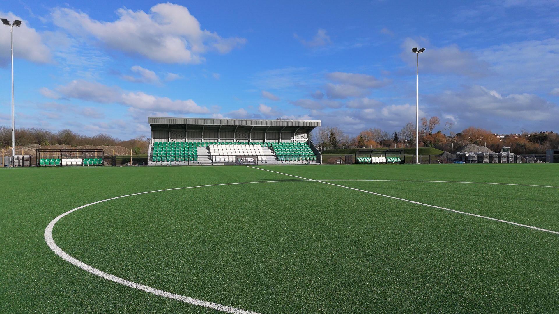 The pitch at Victoria Park is covered with bright green grass and crisp white lines on a bright blue-sky day. There is a stand for spectators with green and white seats. There are floodlights around the pitch.