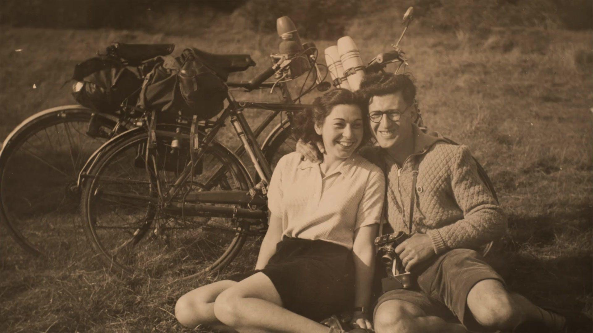 A young couple smile together on a sunny day, sitting in a field, with two bicycles behind them