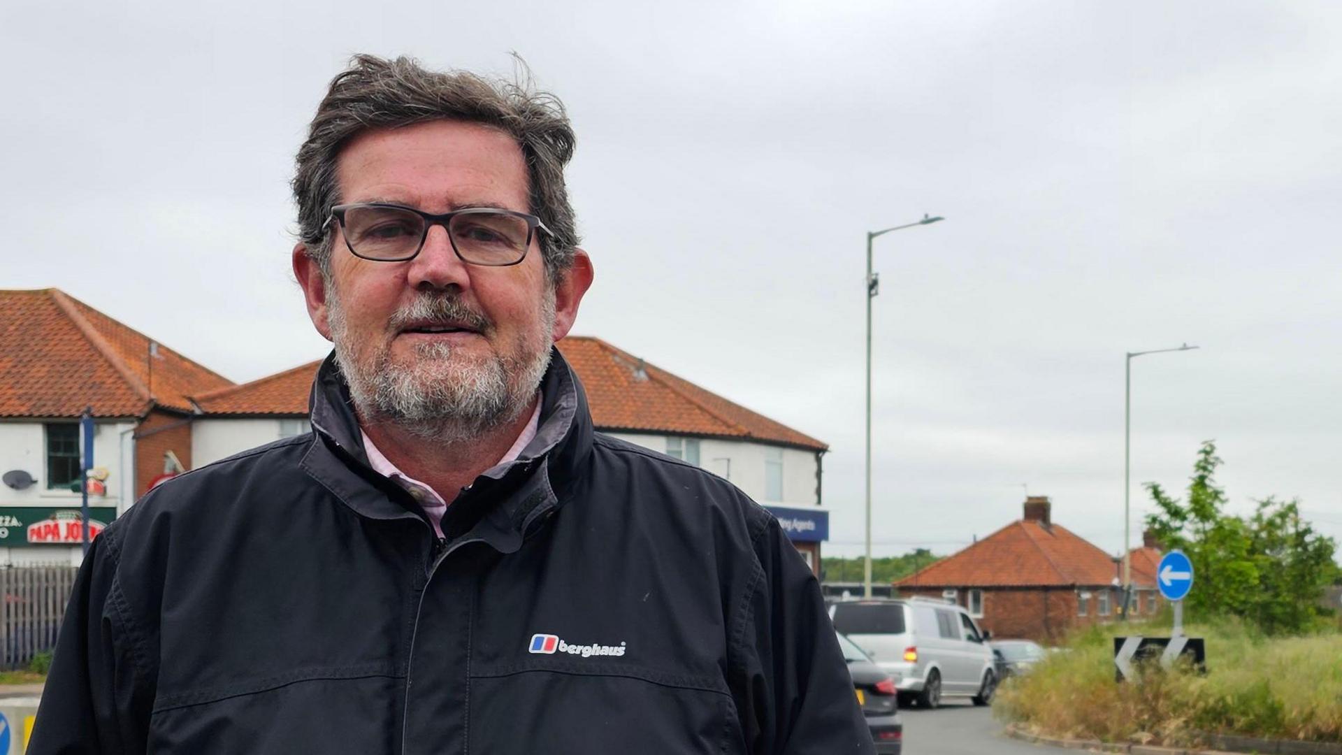We can see Peter Silburn standing on a street with a roundabout in the background. He's wearing a dark blue 'berghaus' jacket and glasses.