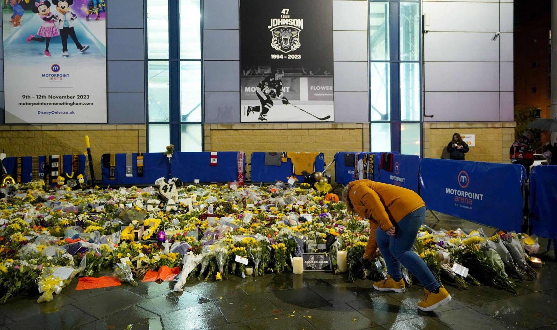 A person adding to a large number of floral tributes, in front of a poster showing Adam Johnson