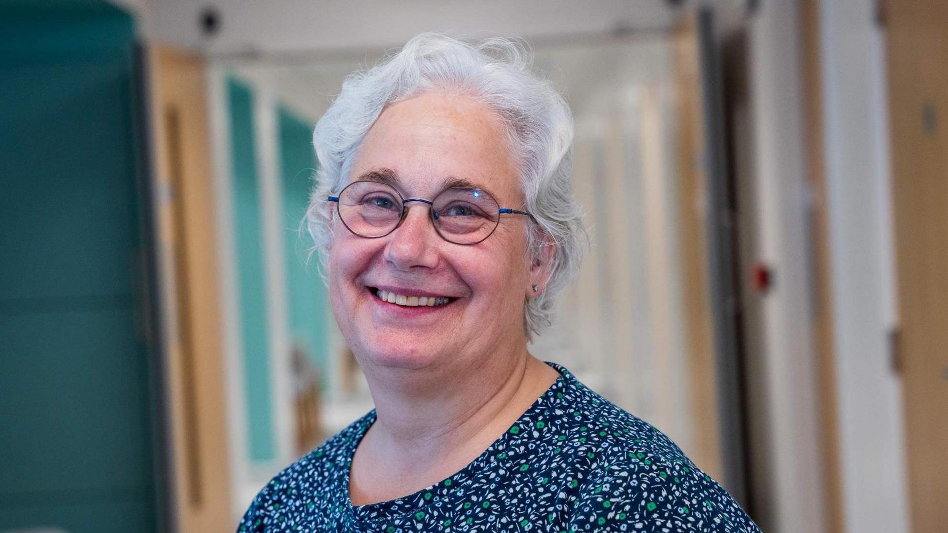 A white-haired woman wearing glasses and a blue top with green and white petal details.  