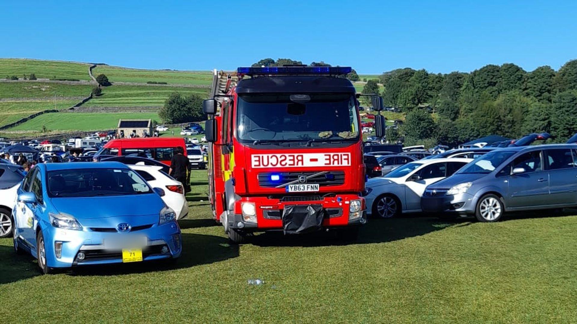 A red fire engine is parked in a field in the middle of a number of parked cars. A row of trees are in the background.