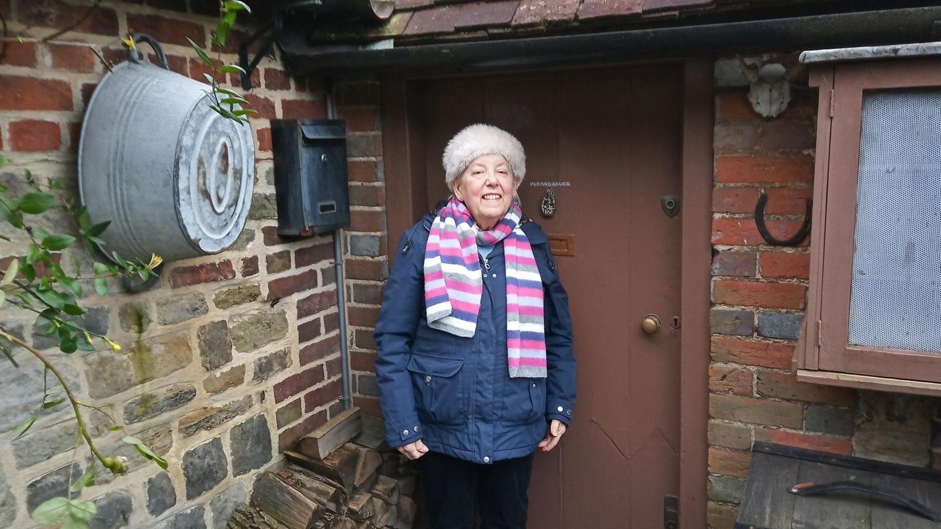 Debby Stevenson, dressed in a blue winter waterproof jacket, striped scarf and white fluffy hat, stands smiling at the door of a stone and red brick cottage with a brown door. Outside are piles of chopped wood and a horseshoe on the wall.