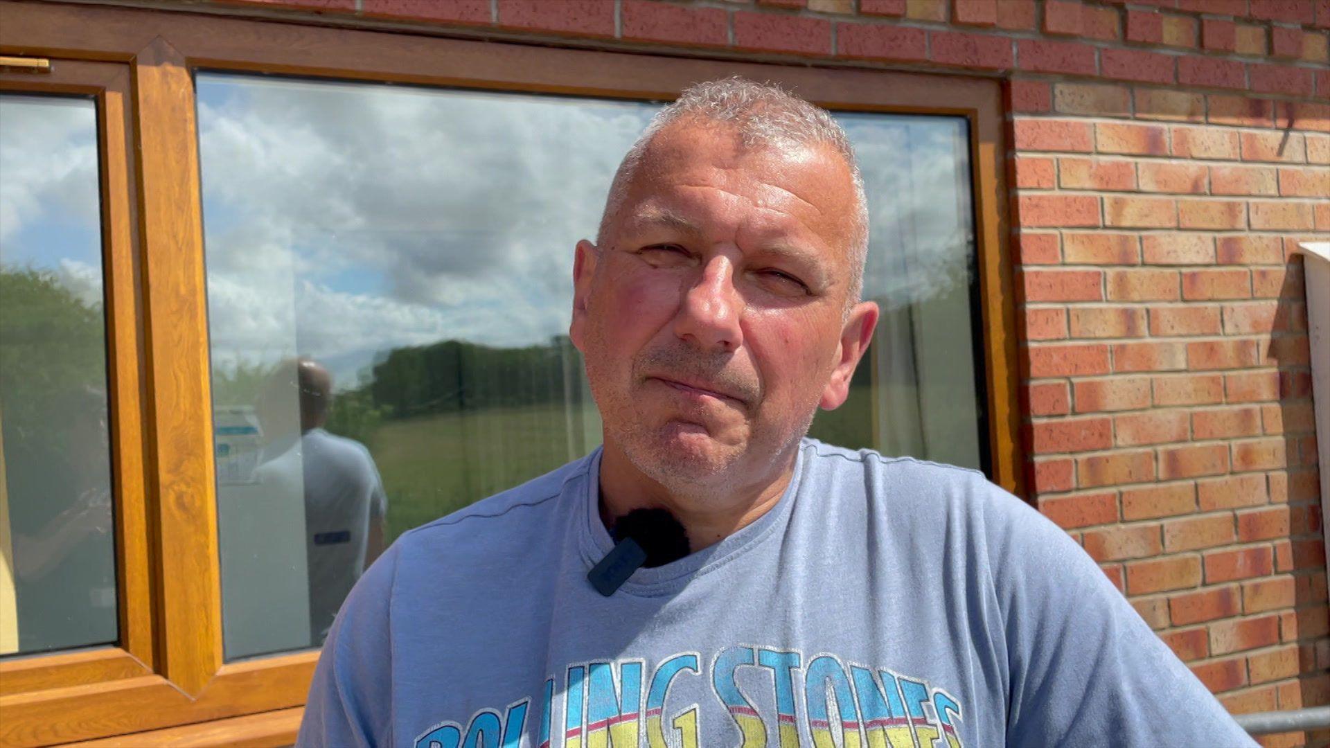A man wearing a purple Rolling Stones t-shirt with a black mini microphone clipped to his collar. He is stood in front of a window of a building and is looking at the camera.