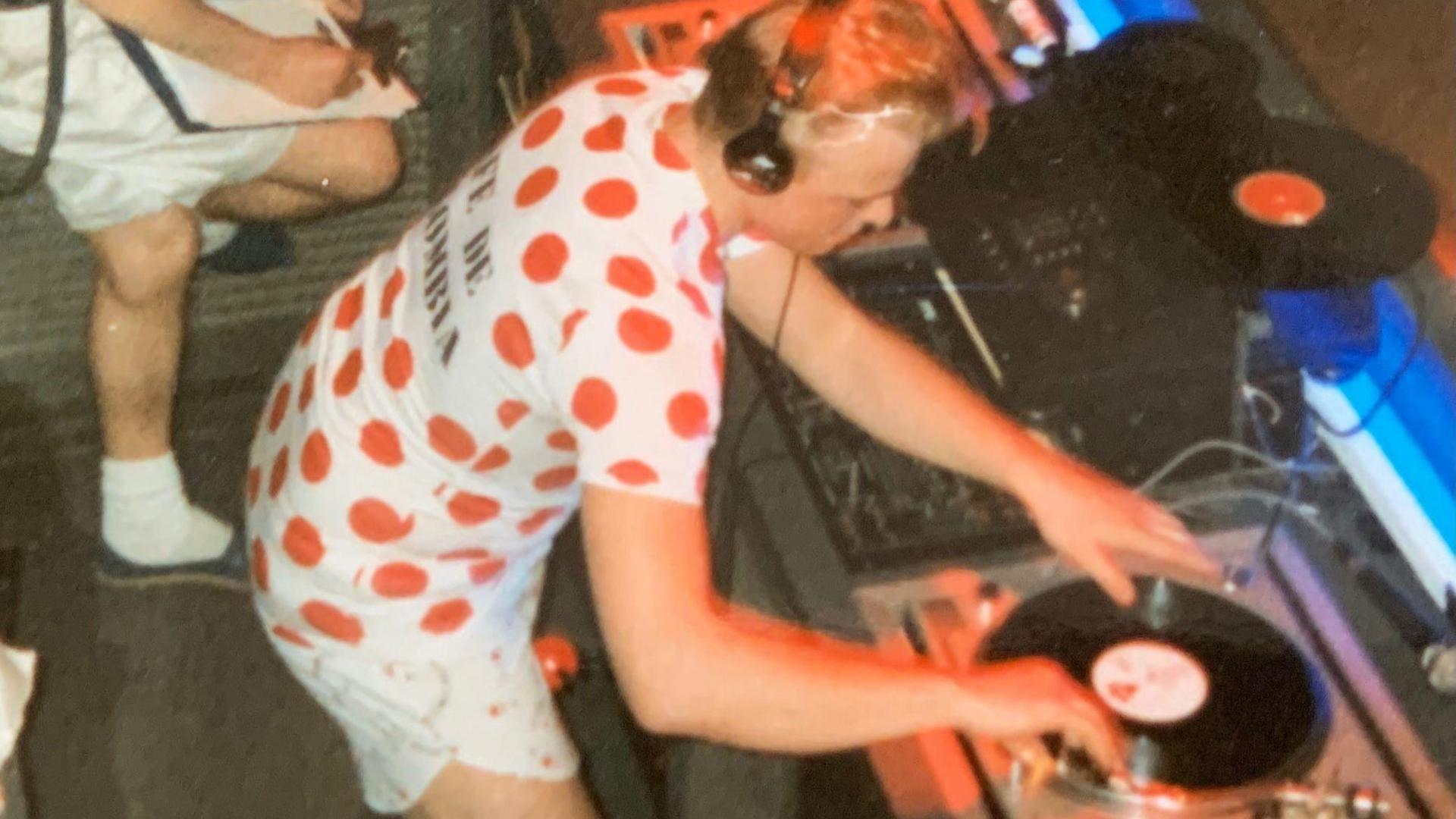 Paul Stainton cues up a record. He is wearing a red polka-dot cycling shirt and shorts. He has blonde hair and is wearing headphones.