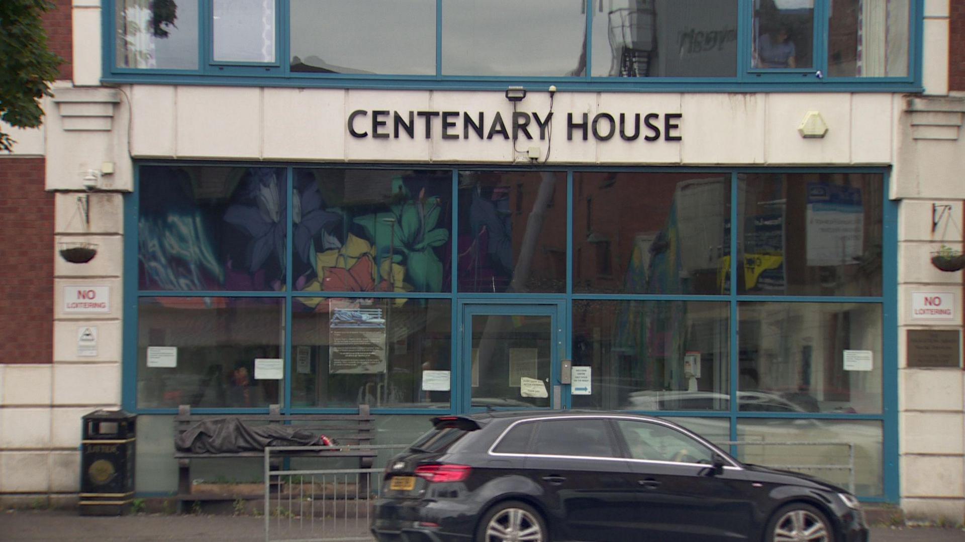 A close up image of the front of Centenary House - the name is spelled out in a metal sign above the entrance which is made up of several plate glass windows and a door. The building is red brick with Portland stone surrounding the entrance. A black Audi is driving past in the foreground.