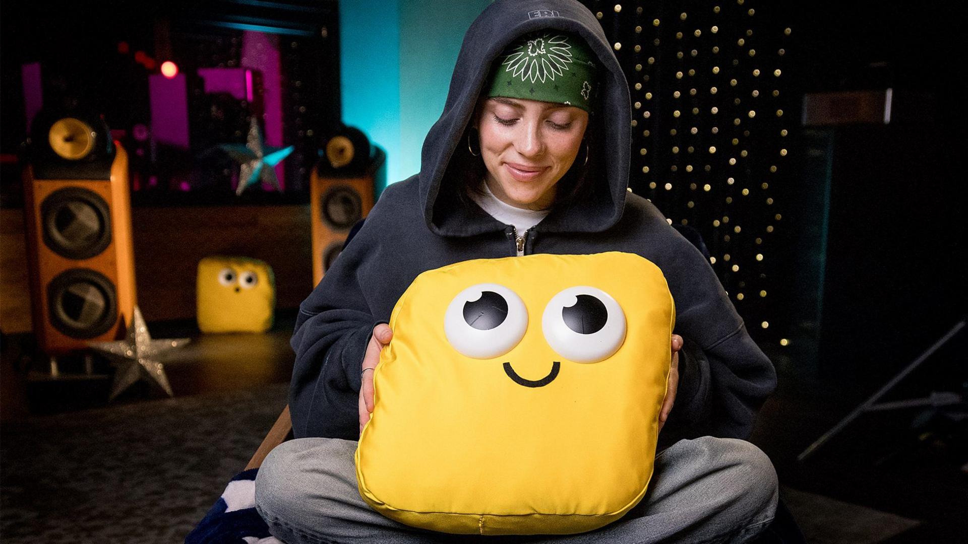 Billie Eilish dark hooded sweatshirt and a bandana is holding yellow cushion with smiley face