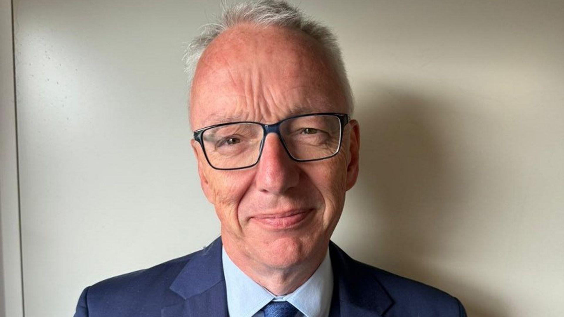 A man with short greying hair, glasses and a blue suit smiling at the camera in an office