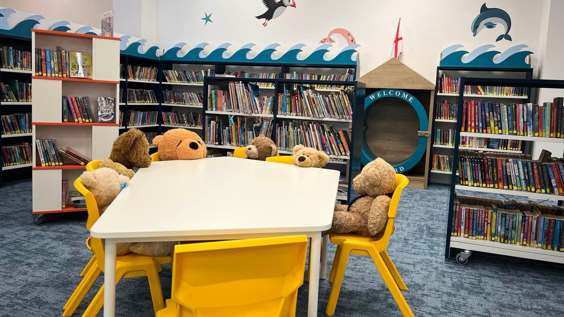 Seven teddy bears sit at a table in Jersey Children's Library. The table is white and the chairs are yellow. Bookshelves with a nautical theme line the walls behind them.