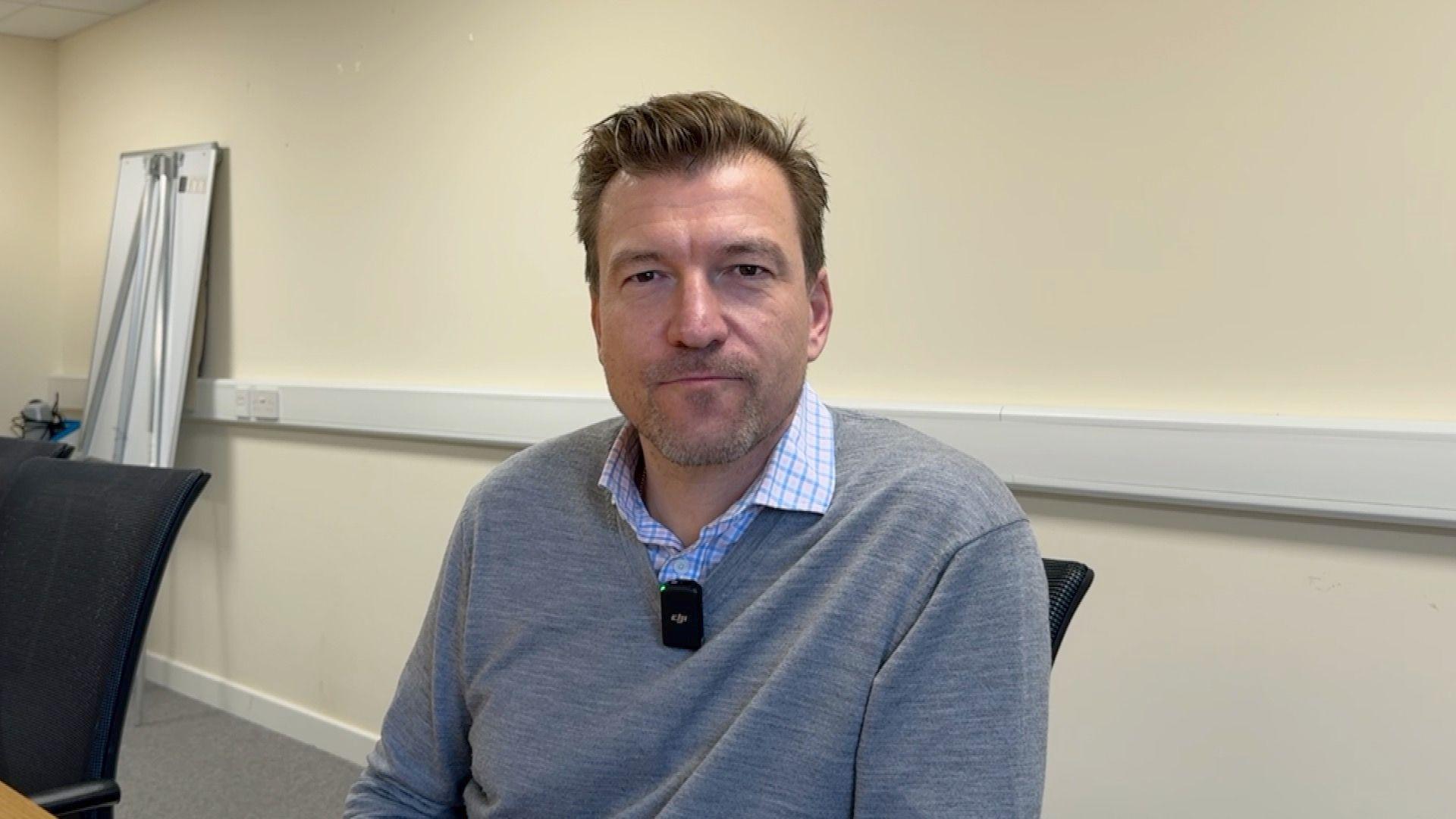 Matt Tabb is a man with brown hair and grey stubble. He is wearing a blue checked shirt and a grey jumper. He is staring at the camera. He is sat in a black chair in a meeting room.