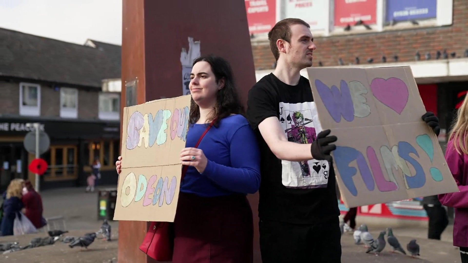 A man, wearing a black t-shirt with the Batman villain The Joker on it, and a woman in a blue jumper and red dress, stand in Kettering town centre holding cardboard signs which support the Odeon cinema. 