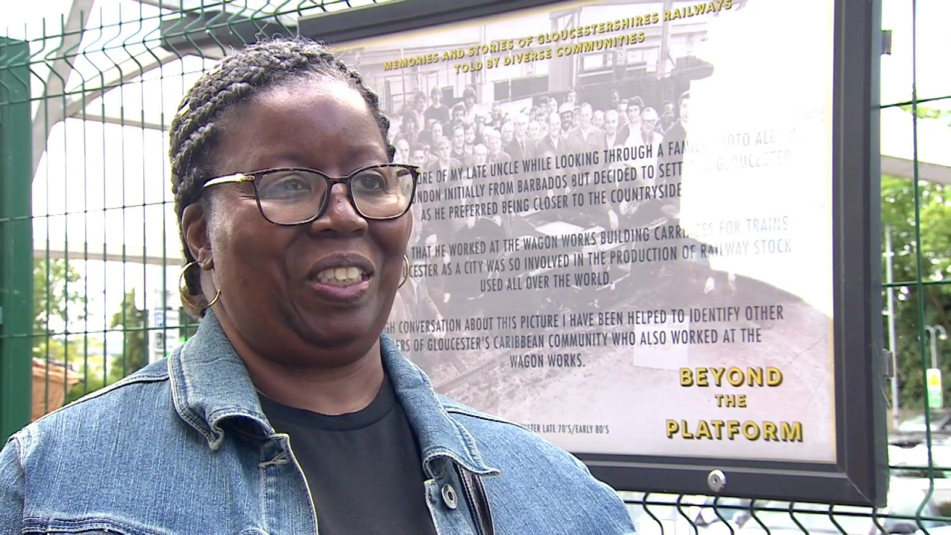 Hazel Golden wearing glasses and a denim jacket standing in front of a billboard for Beyond the Platform.