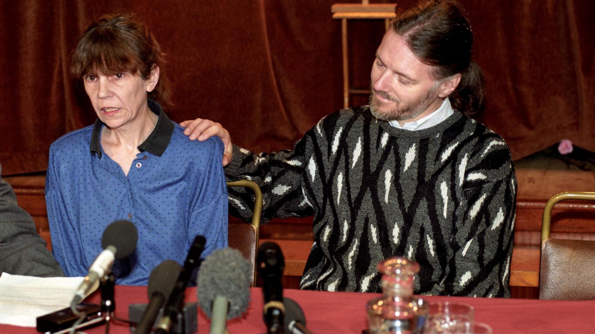 A picture of Geri Rimer with dark hair pulled back alongside her husband Gordon Rimer with a beard and long hair tied in a ponytail. The picture taken in 1994 shows them speaking at a police press conference.