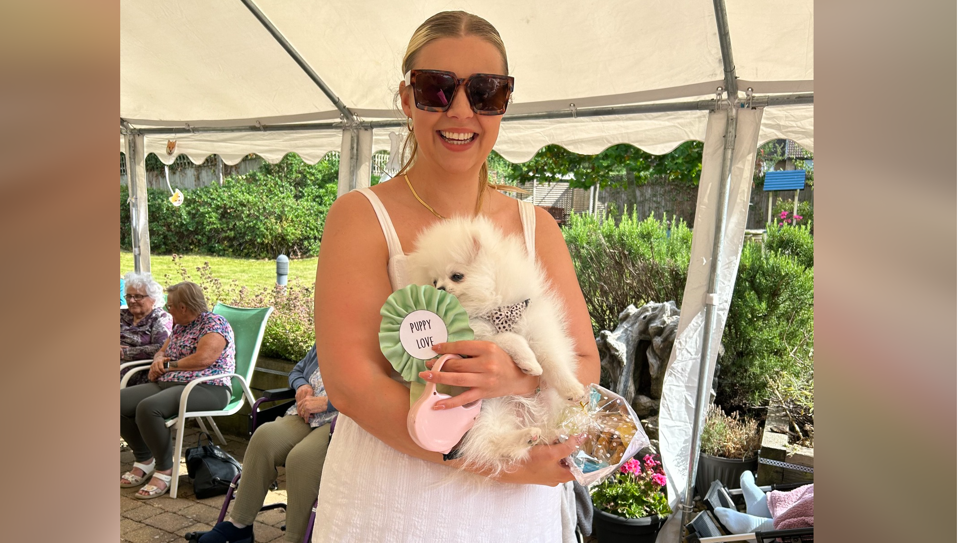 A woman in a white dress holding a white dog with a green rosette which says "Puppy love"