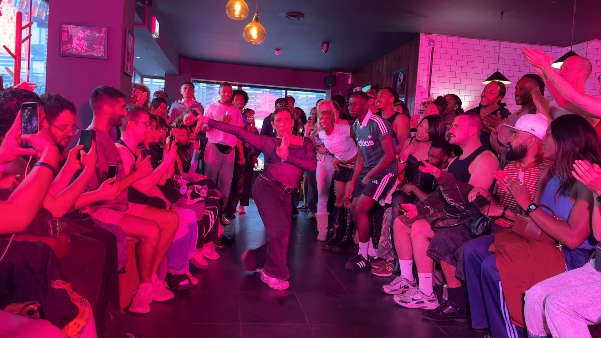 A voguer walks the runway during the event, while a crowd surrounds the floor, cheering and recording on their phones.