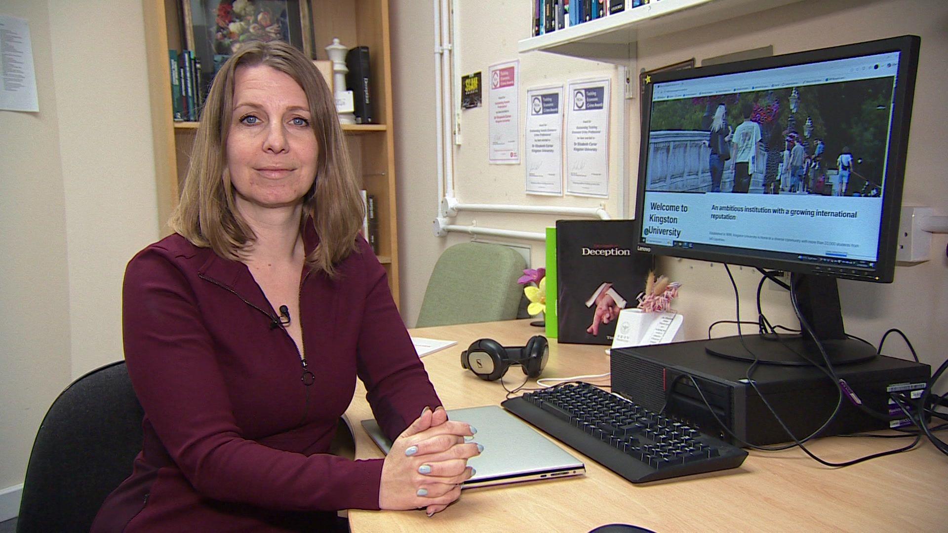 A woman with shoulder length brown hair sits at a desk and faces the camera. She rests her hands together on a desk in front of her. She is wearing a dark red dress. A computer screen is on her desk as well as a laptop that is closed, headphones and other office items.