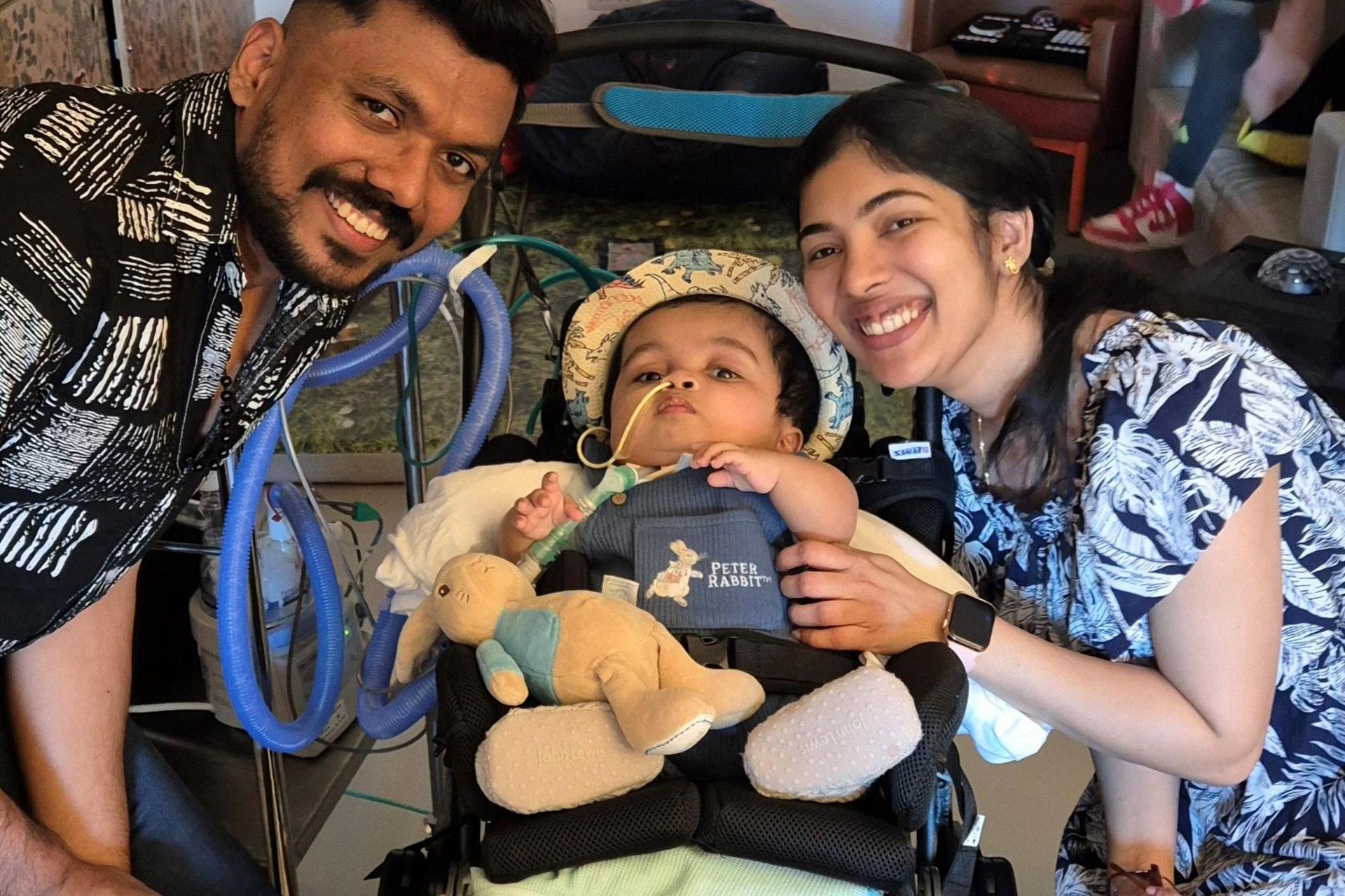 Gabriel is in a special pushchair that has a ventilator attached. He is wearing a hat and has a rabbit teddy. His parents flank him and are smiling at the camera. His dad is wearing a black and white shirt and his mum is wearing a black and white dress.