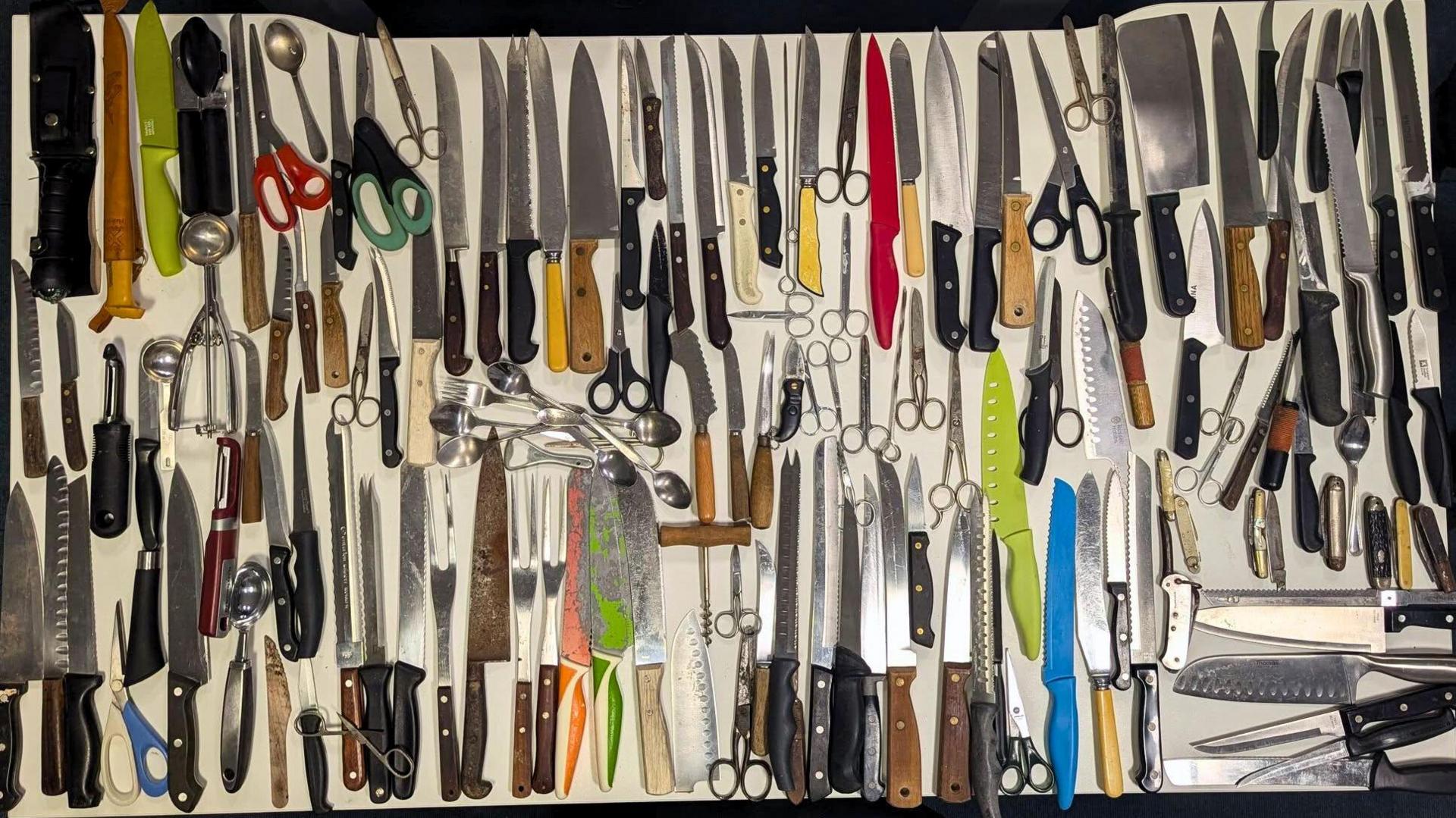 A large assortment of kitchen knives and utensils is neatly arranged on a white surface. The collection includes chef’s knives, cleavers, serrated knives, paring knives, utility knives, and multiple pairs of scissors. Also visible are sharpening steels, peelers, can openers, ice cream scoops and spoons.