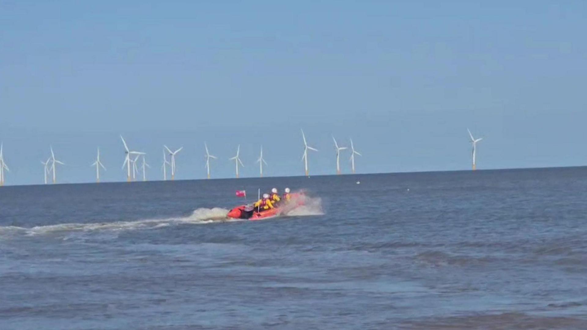An inshore lifeboat - a small, orange craft carrying three people wearing yellow jackets, red lifejackets and white helmets - makes waves as it heads out into a calm-looking blue sea under a blue sky, with white wind turbines on the horizon.