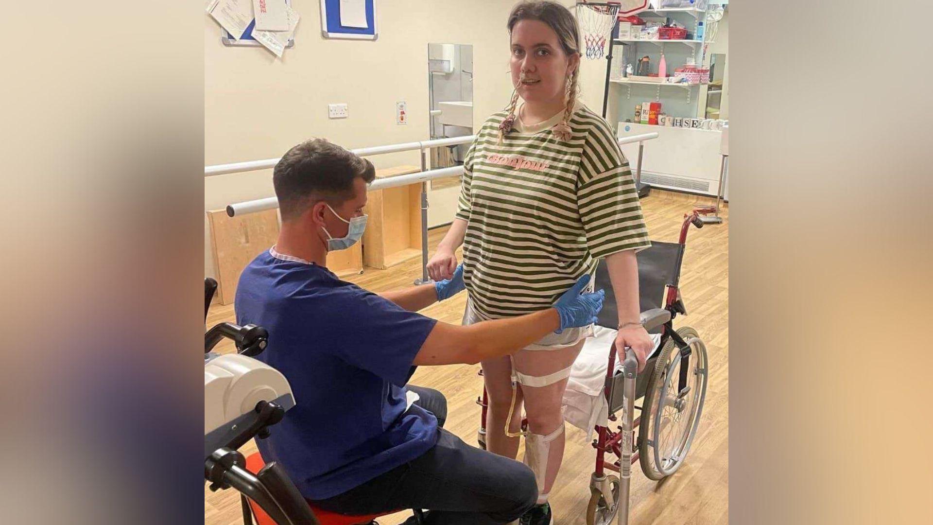 Ellen has her hair in pig tails and is stood up from a wheelchair. She has a stripy t-shirt and has medical tape on her left leg. A physiotherapist is sat on a chair in front of her with his arms out helping her to stand