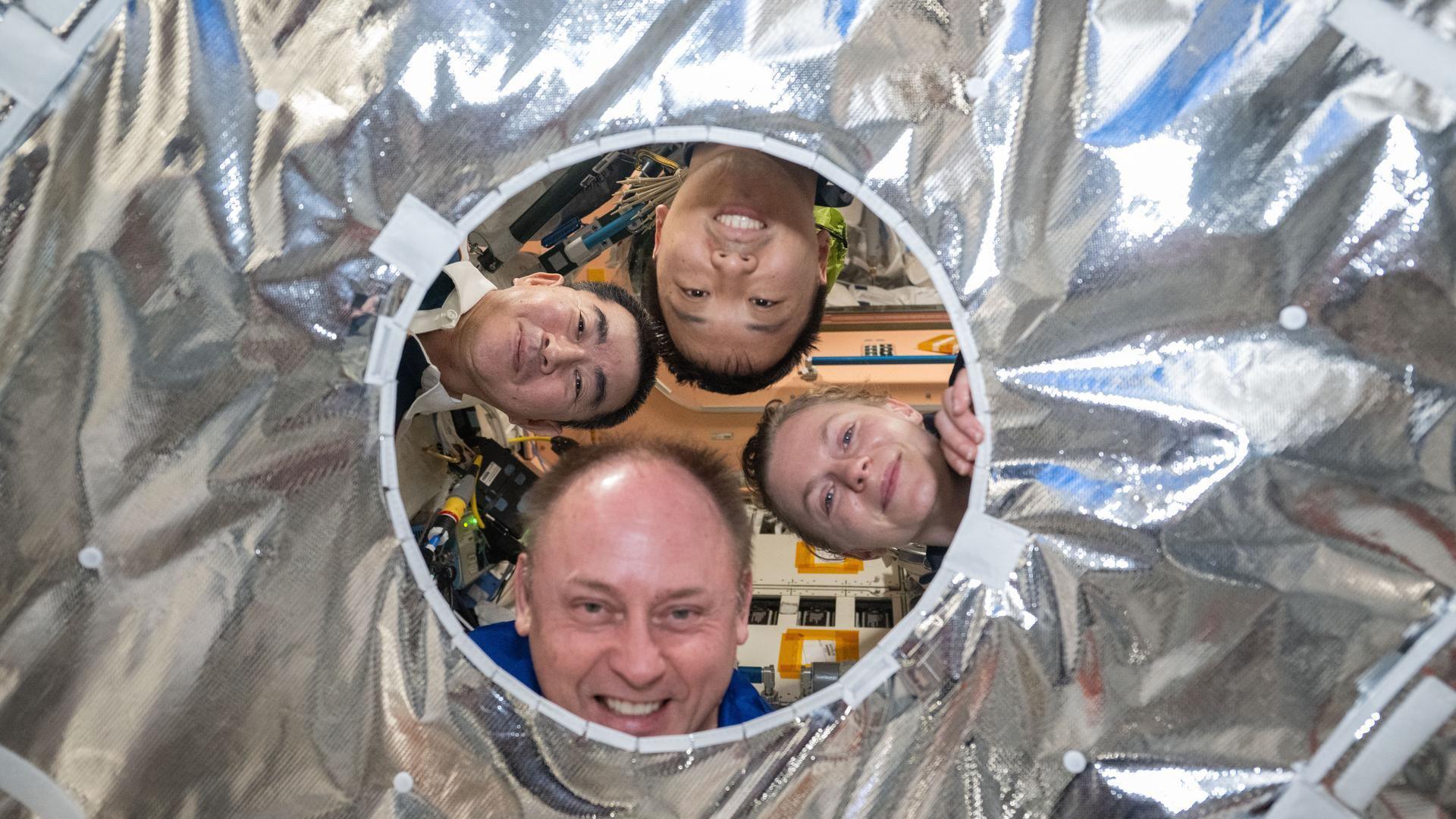 Four ISS crewmates peer through a circular opening in a hatch thermal cover aboard the International Space Station. Clockwise from left, are Kimiya Yui, Jonny Kim, Zena Cardman, and Mike Finck