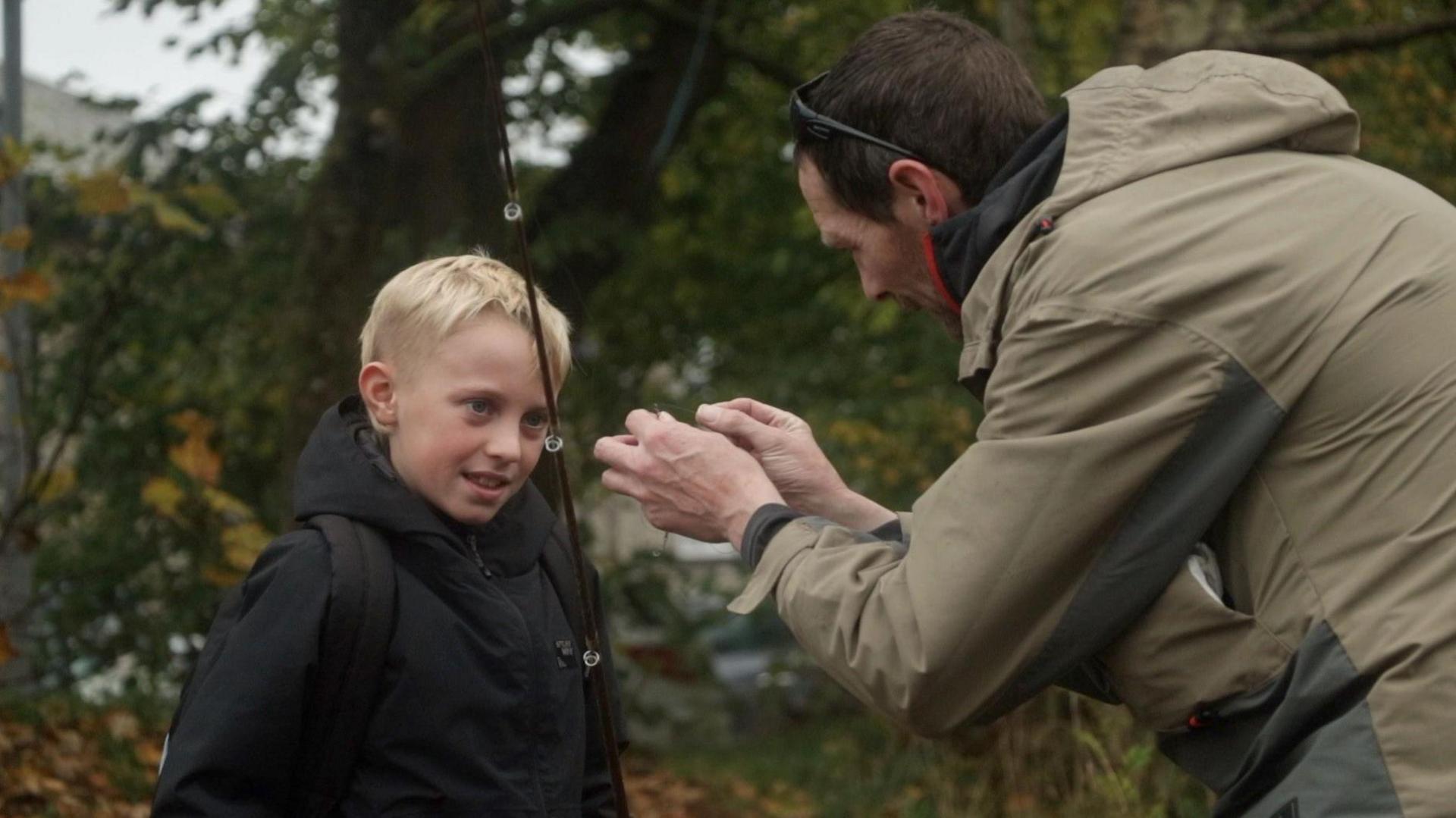 Fisherman Kieron Banks prepares a rod and a line while a young boy, who has blonde hair, looks at the hook being prepared. They are standing by a river bank which has lots of green shrubbery. 