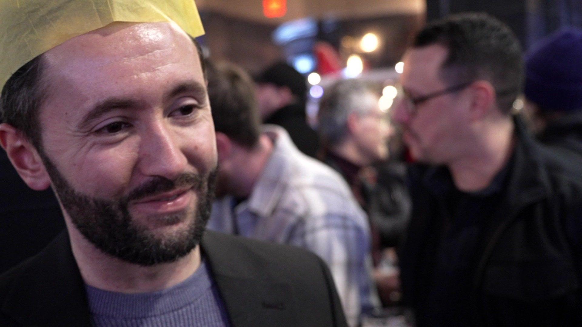 Man in blue jumper and black blazer wearing a yellow paper Christmas hat stands outside a pub