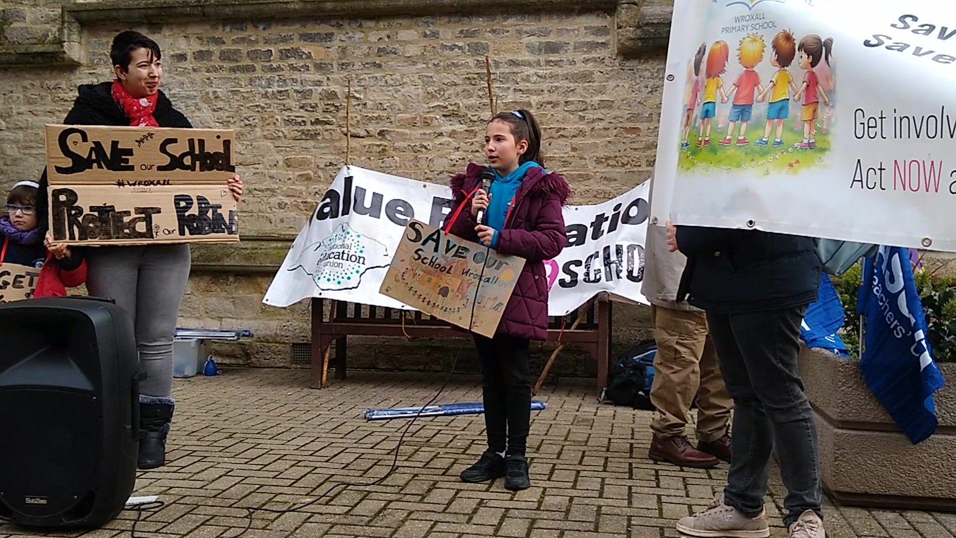 A young girl stood holding a cardboard sign and a microphone. Next to her are three other people who are holding banners. All of the signs say "save our schools".