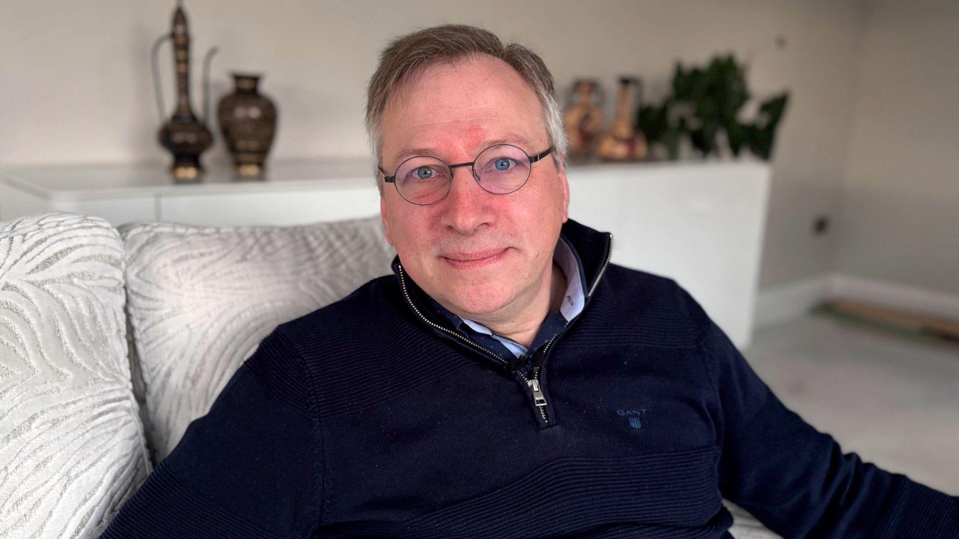 A man with round glasses and short grey hair sits on a silver sofa, looking down the lens of the camera. He has his lips pursed and his eyes are a piercing blue colour. He is wearing a navy quarter zip jumper with a silver zip.