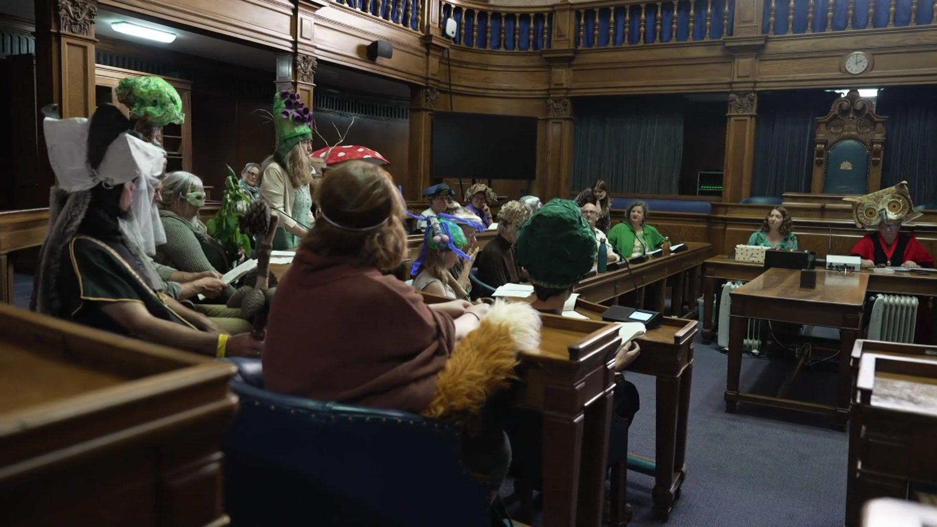 A group of people sat in a council room dressed in various nature inspired costumes, including a tree