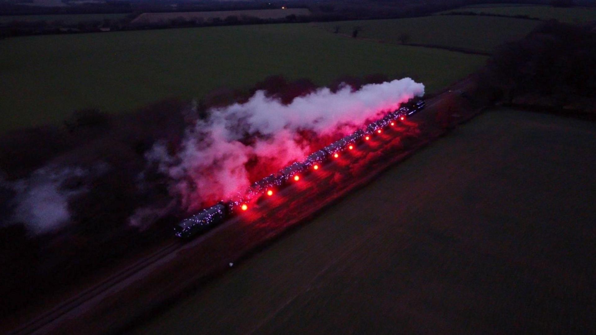 An aerial view of the train travelling through the countryside at night. Steam puffs out of the train, which is adorned by red lights on its side.