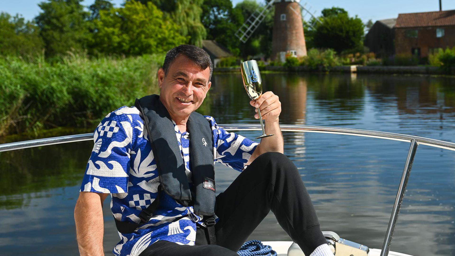 Mr Harvey is sitting on the front of a white boat which is travelling on a small river. He is wearing a blue and white shirt and a black life jacket, and is holding a gold champagne glass in the air. 