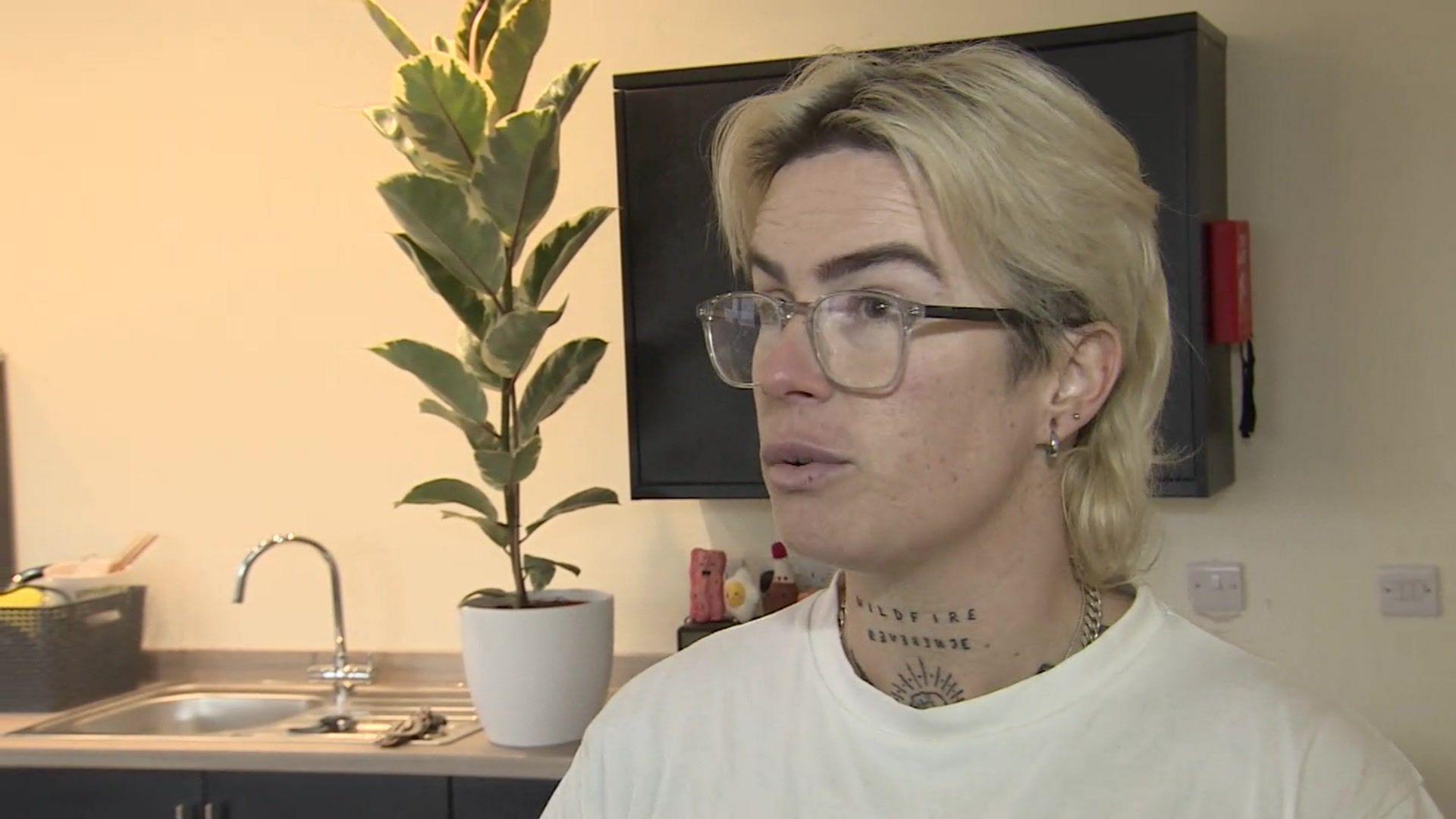 Ren in her new flat in Maryport being interviewed by BBC Look North. She has short blonde hair and wears glasses. She is looking away from the camera, she is wearing a white top and has a tattoo on her neck. Behind her is a kitchen sink, a plant and a TV.