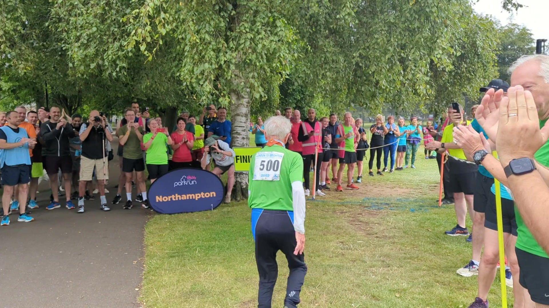 Parkrun runners stand in two lines cheering as Bob Emmerson runs through the crowd. He is wearing a green running shirt with the number 500 on the back. 