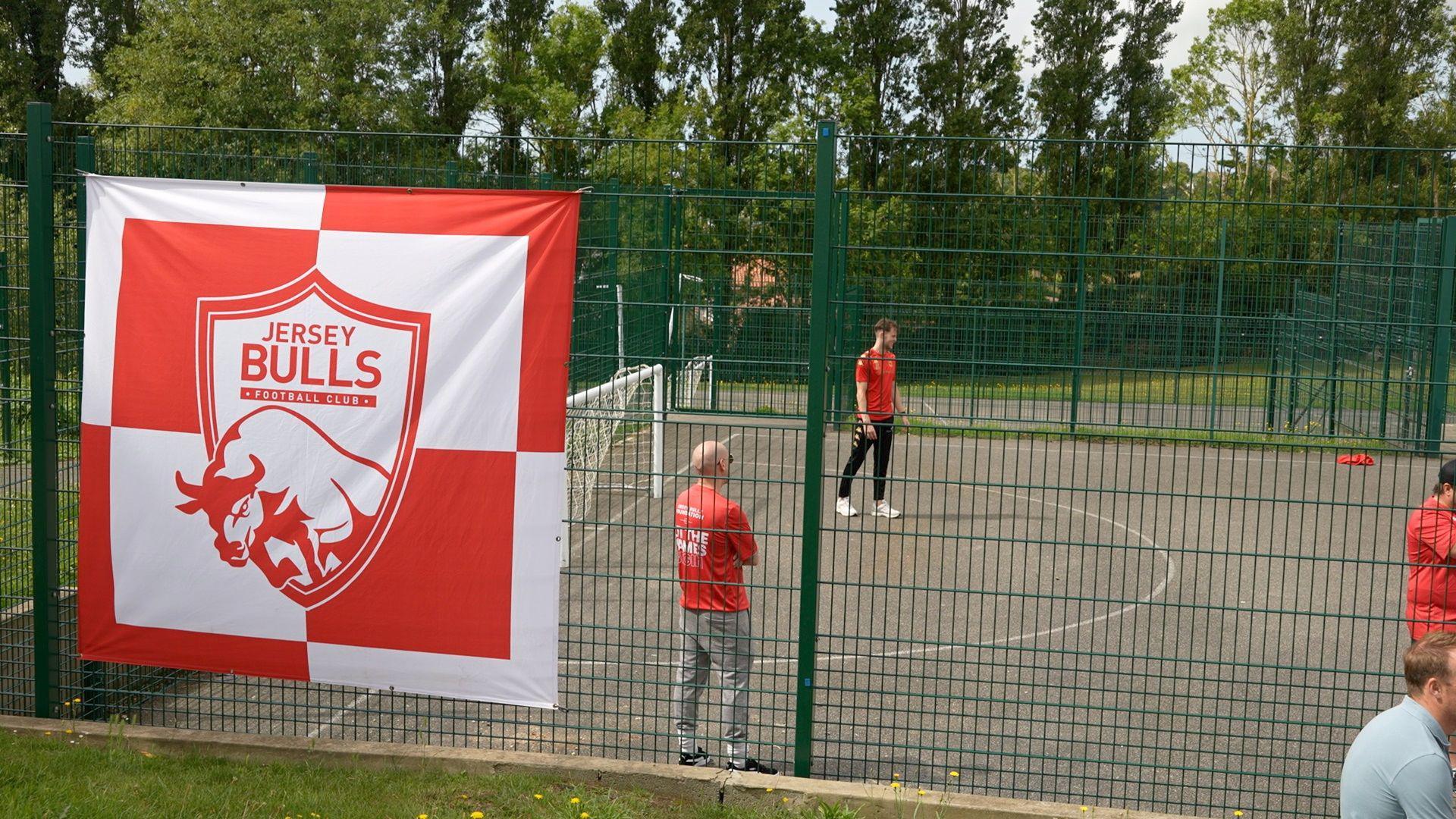 There's a flag with the Jersey Bulls logo on and the flag is attached to a metal fence which is around a concrete five-a-side football pitch with two men inside who are in the red Jersey bulls tops.