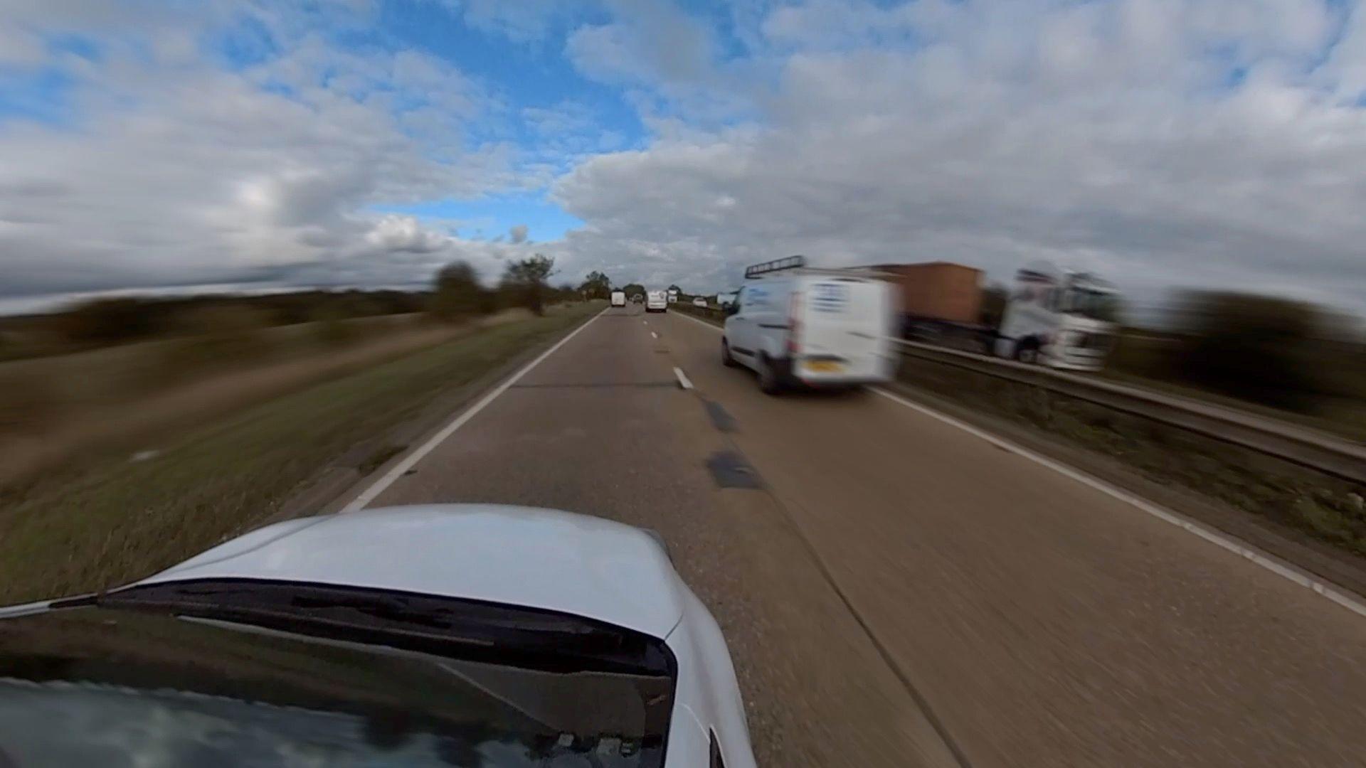 The view of driving down the A12 fro a white car. A van is over taking on the right. Open fields sit on the left.