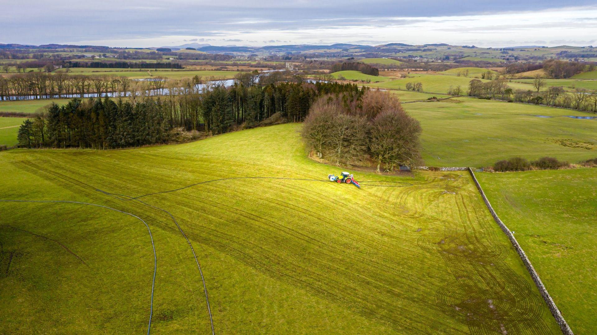 An aerial view of a tractor applying slurry to fields, in the background are a rolling patchwork of fields and a river winding through some evergreen forest. The sky is a cloudy and grey but brighter on the horizon. 