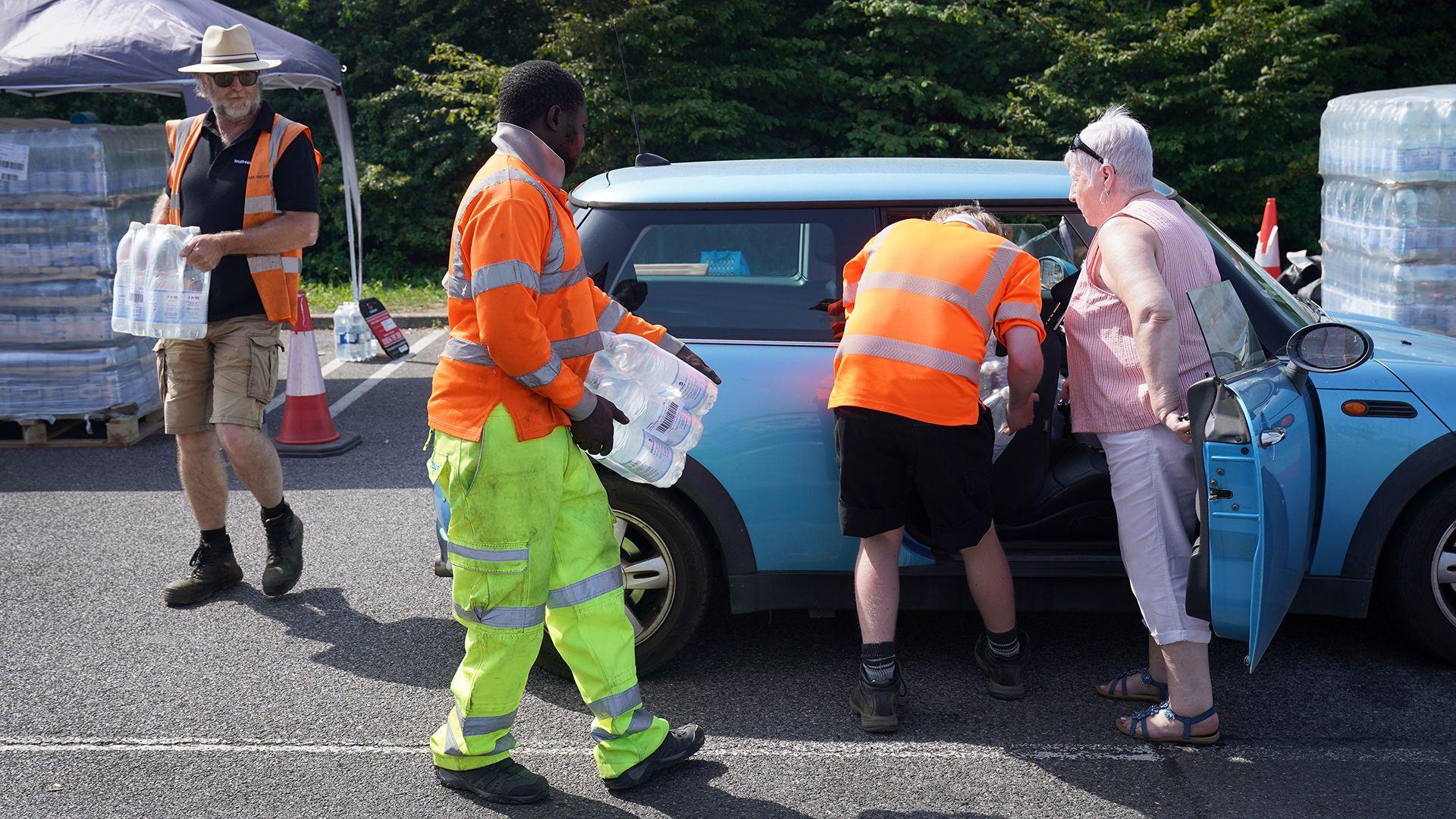 People in high vis uniforms distribute bottled water to a woman's car on a road in Herne Bay. There's a gazebo and stacks of water in the background and trees line the road.
