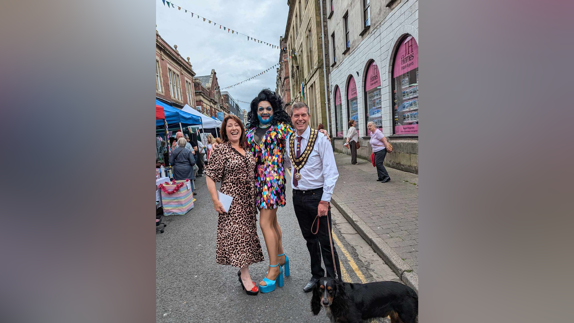 The three are standing in the street with stalls on one side. Patty is wearing a sparkly multi-coloured dress, the town mayor is wearing ceremonial medals and he is holding a dog. Ms Scrogham is wearing a leopard print dress. All are smiling.