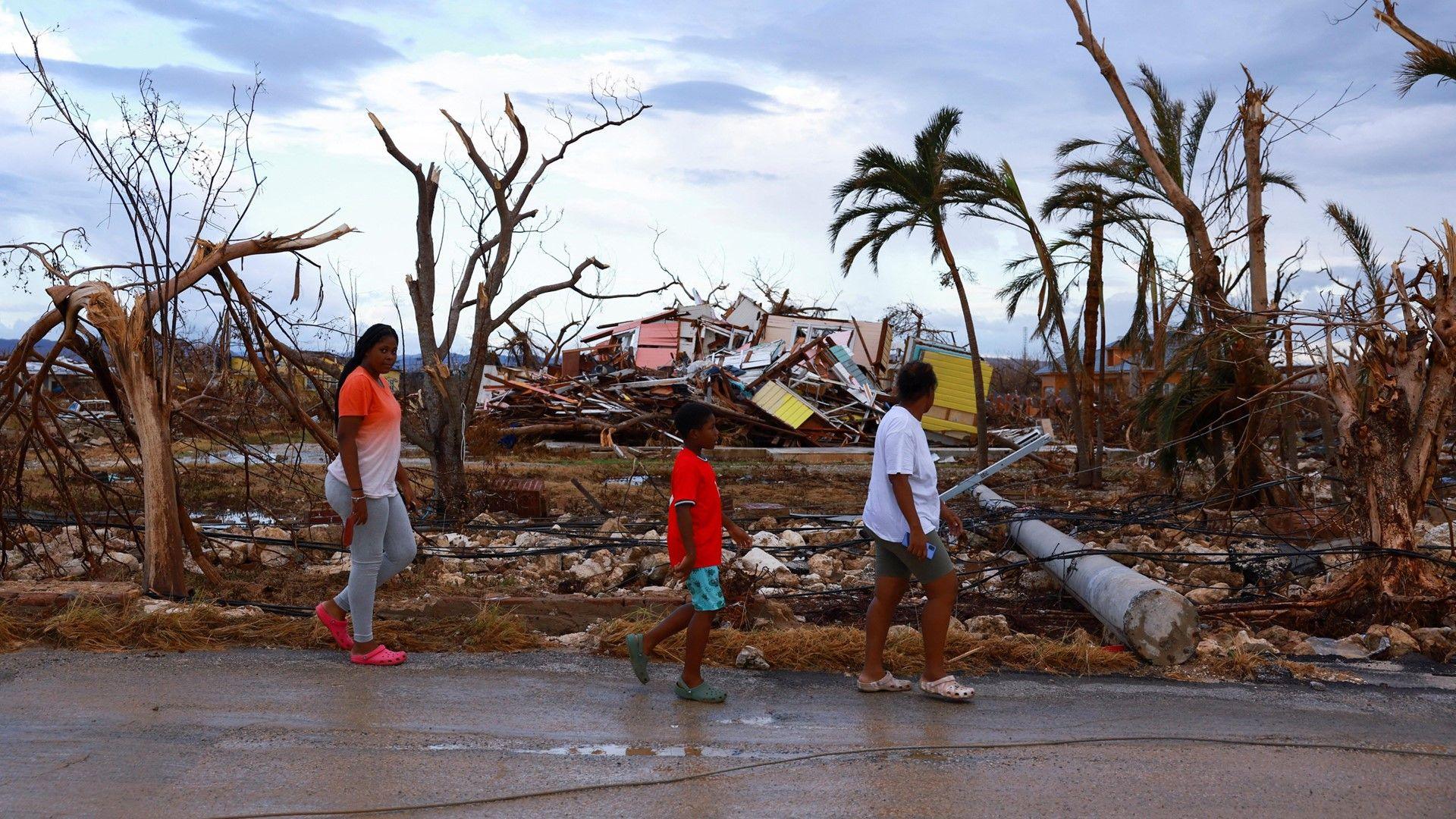 Three people walking past broken trees and blown down houses left by the passage of hurricane Melissa.